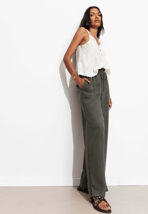 Femme aux longs cheveux portant un haut blanc sans manches, un pantalon ample vert olive et des sandales noires cloutées, debout contre un mur blanc.