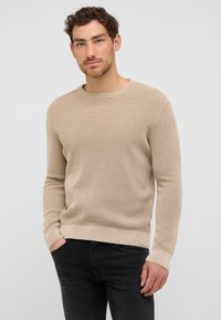 Beżowy sweter dziany z okrągłym dekoltem i długimi rękawami, z teksturowanym wzorem. W połączeniu z ciemnymi dżinsami, tworzy luźny wygląd.