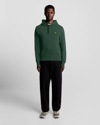 Haut à capuche vert avec une poche avant et un cordon de serrage, associé à un pantalon noir ample et des baskets grises avec des accents blancs.
