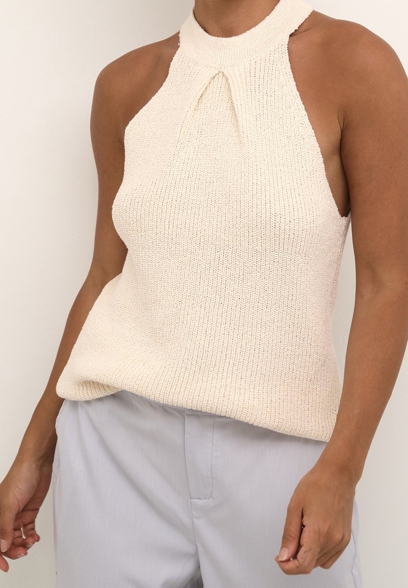 Haut dos-nu en maille beige clair avec une découpe en forme de trou de serrure. Il présente un col montant, une texture côtelée et une coupe ample, associé à un pantalon gris clair.