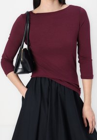 Burgundy långärmad topp med båturringning, tillsammans med en svart plisserad kjol, kompletterad med en liten svart väska med gulddetaljer på dragkedjan.