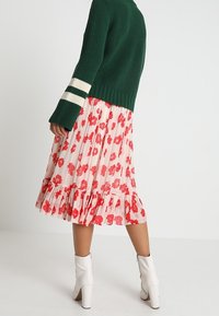 Maglione verde a coste con strisce crema sulle maniche, abbinato a una gonna rosa con motivi floreali rossi e bordo arricciato. Stivaletti bianchi alla caviglia.