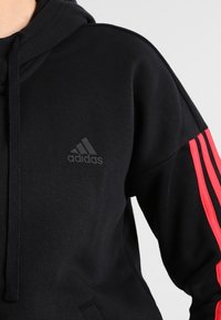 Sweat à capuche noir en tissu à finition texturée. Présente des rayures rouges sur les manches et un logo ton sur ton sur la poitrine.