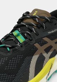 Chaussure de sport noire en tissu texturé, avec des accents marron et un motif en zigzag sur le côté. Présente une semelle intermédiaire jaune et des détails verts sur les lacets.