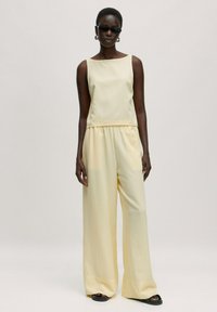 Helles Gelb, ärmelloses Oberteil mit geradem Schnitt, kombiniert mit weiten Hosen aus passendem Stoff. Glatte Textur, minimale Details, entspannter Schnitt.