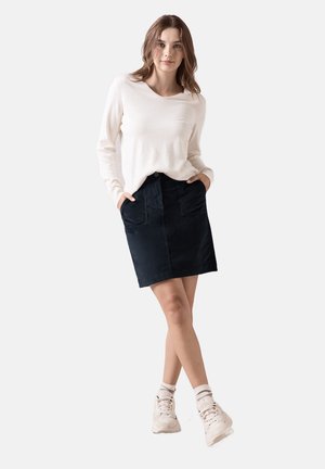 Jupe en denim bleu marine avec deux poches frontales, associée à un pull léger crème. Le modèle porte des baskets blanches et des chaussettes retroussées.