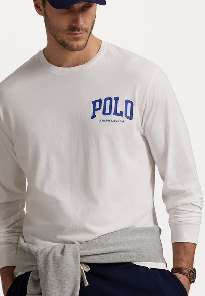 Ilga rankovė, balta, medvilninė marškinėlių su mėlynu "POLO" logotipu. Pilkas džemperis, surištas aplink liemenį. Paprastas dizainas, laisvas kirptis, klasikinis stilius.