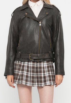 Frau trägt eine dunkelbraune Leder-Bikerjacke mit Gürtel über einem weißen Hemd und einem braunen karierten, plissierten Rock vor weißem Hintergrund.