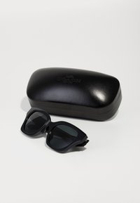 Lunettes de soleil noires avec verres foncés et design de monture géométrique, à côté d'un étui rigide noir arborant un logo en relief sur la surface.