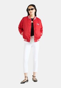Veste bomber en satin rouge avec des poignets et un col à rayures blanches, portée par-dessus un haut noir et un pantalon blanc court, complétée par des ballerines noires.