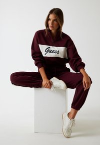 Bordeaux zip-up sweatshirt met wit paneel met de tekst "Guess" in marineblauw. Gecombineerd met bijpassende joggingsbroeken en witte sneakers. Soepele textuur, ontspannen pasvorm.