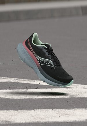 Juoda ir mėtų spalvos bėgimo batelis su koralinės ir pilkos spalvos padu, ore virš asfalto su baltais linijų žymėjimais.