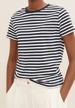 Person trägt ein marineblau-weiß horizontal gestreiftes Kurzarmshirt, das in hoch taillierte weiße Hosen mit Knopfverschluss gesteckt ist.