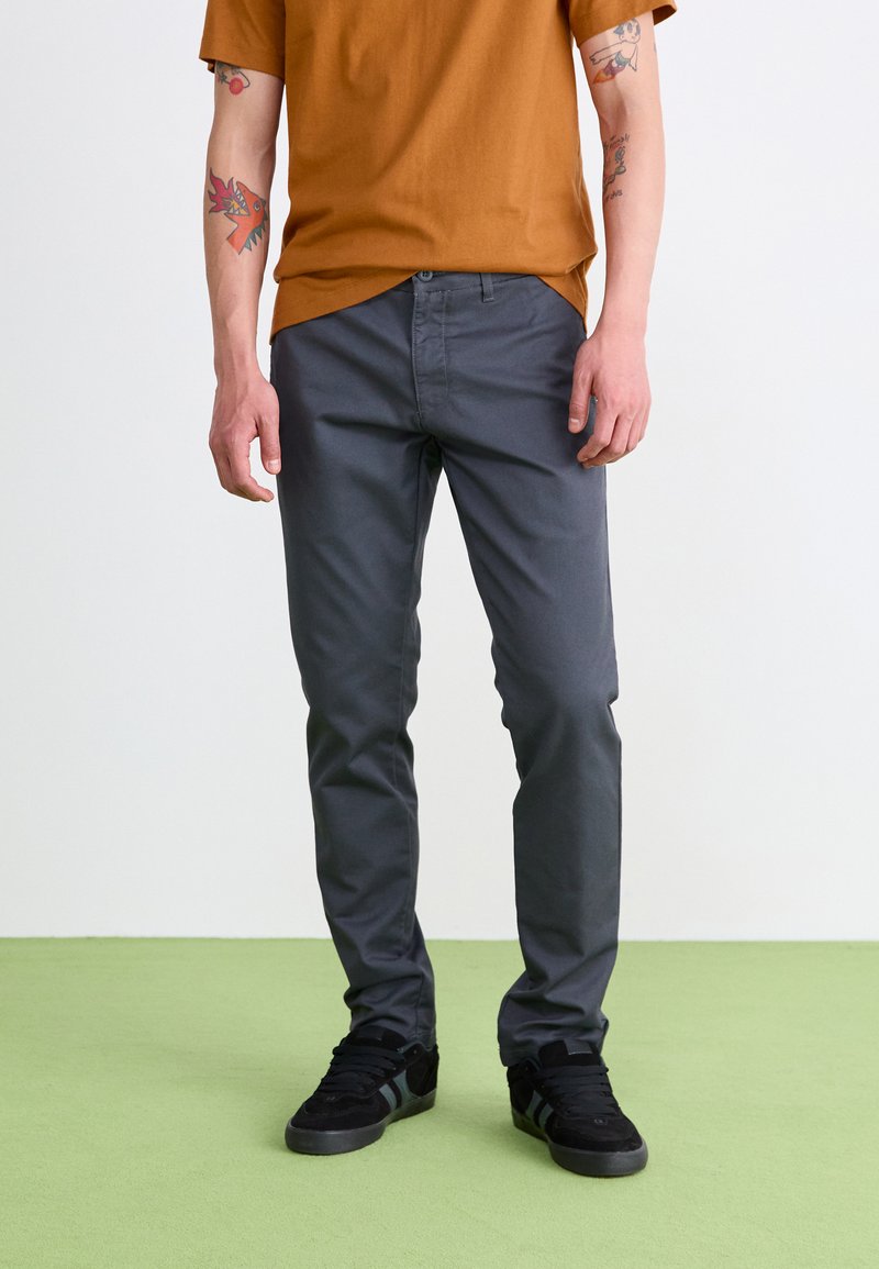Graue Baumwollhosen mit schmaler Passform, Knopfverschluss, seitlichen Taschen und Details an der Gesäßtasche, kombiniert mit einem rostfarbenen T-Shirt und schwarzen Sneakers.