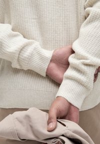 Ljus crème-färgad stickad tröja med ribbade muddar, bärs över lätt beige tyg. En hand håller ett vikt ljusbeige plagg.