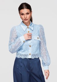 Femme portant un chemisier en dentelle bleu clair avec des boutons dorés et un col en satin, associé à un pantalon taille haute bleu foncé, posant devant un fond uni.