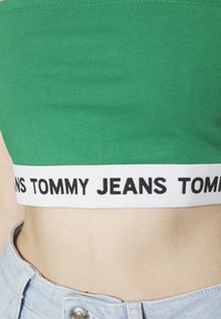 Zelený cropped top s bílým elastickým páskem s nápisem "TOMMY JEANS" v černém písmu. Hladká textura, přiléhavý střih. Kombinováno s džíny.