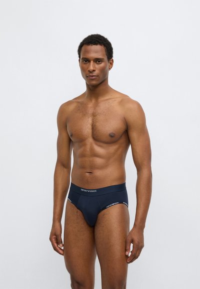 Zilas kokvilnas biksītes ar logotipu jostasvietā. Īpaši pieguļošs dizains, gluda tekstūra, minimālas šuves. Modelis demonstrē produktu bez papildu apģērba.