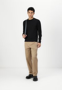 Black long-sleeve t-shirt, beige chinos, and black shoes. Standing pose with a gray strap across the shoulder. Simple design, no patterns.