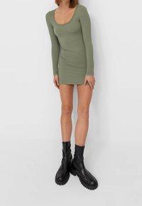 Robe à manches longues côtelée vert olive avec un décolleté en U, associée à des bottines noires chunky. La robe est ajustée et de longueur mini.