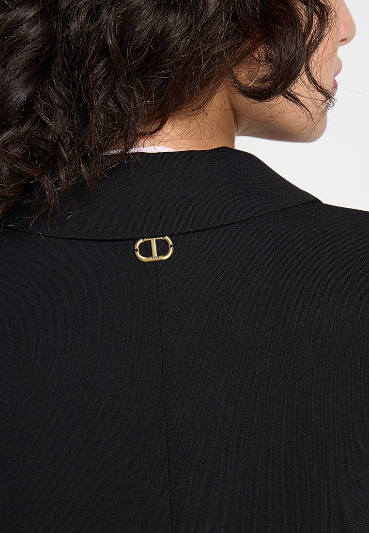 Blazer noir ajusté en tissu texturé subtil, avec une fermeture dorée à l'encolure arrière.