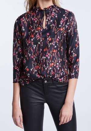 Frau trägt eine schwarze Bluse mit kleinen rosa und roten Blumen und schwarzen hoch taillierten Hosen, steht mit entspannten Armen.