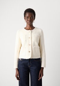 White cropped cardigan in soft, textured fabric with fringe trim, featuring four patch pockets and decorative buttons. Paired with blue jeans.