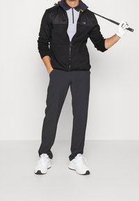 Mężczyzna w czarnej kurtce, szarym polo i ciemnych spodniach trzymający kij golfowy na ramieniu, w białych rękawiczkach i białych trampkach.