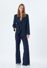Blazer blu a righe sottili con revers ampio, chiusura a un bottone e due tasche, abbinato a pantaloni blu e scarpe appuntite argentate.