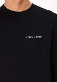 Čierny bavlnený sveter s okrúhlym výstrihom a bielou vyšívanou značkou "LYLE & SCOTT" na ľavej hrudi. Hladká textúra.