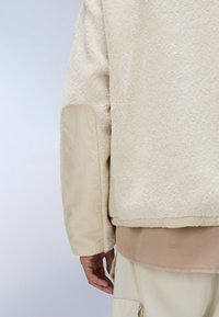 Jachetă din fleece de culoare crem, cu un exterior texturat și moale. Prezintă un panou bej contrastant pe mânecă și o margine cu nervuri. Pur și simplu peste o cămașă bej.