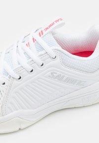 Scarpa sportiva bianca con tomaia texturizzata, linguetta in rete e dettagli rosa. Presenta un logo prominente e un design della suola a contrasto.