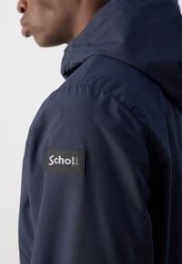 Marineblå jakke med teksturert overflate, med et Schott-logo på skulderen. Designet inkluderer hette og rene linjer.