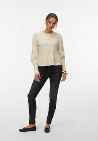 Femme portant un cardigan crème boutonné, un jean skinny noir, des ballerines noires et un bracelet, debout avec une jambe légèrement pliée.