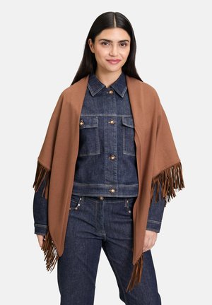 Femme portant une veste en denim foncé et un jean, avec un châle à franges marron drapé sur ses épaules, se tenant devant un fond blanc.