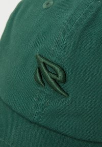 Green cotton baseball cap featuring a textured surface, embroidered logo in a matching shade, and a curved brim.