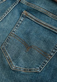 Buzunar din denim cu spălare deschisă, cusături vizibile în portocaliu și o formă rectangulară clasică. Textură netedă, aspect ușor uzat.