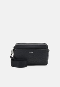 Black crossbody bag with textured logo pattern, zip closure, smooth material, and adjustable strap. Features subtle branding on the front.