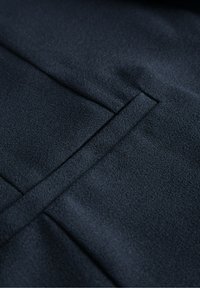 Dunkelblauer Stoff mit glatter Textur, der eine Naht und eine Taschenanzeige aufweist. Die Oberfläche zeigt einen subtilen Glanz und eine feine Struktur.