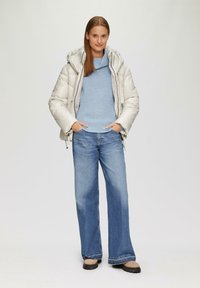 Hellgraue Daunenjacke mit Kapuze, über einem blauen Strickpullover getragen. Lockere blaue Jeans mit geradem Bein und schwarzen Schuhsohlen.
