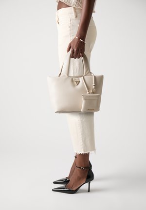 Beige syntetisk tote bag med doble håndtak, gulllogo detaljer og avtagbar lomme; kombinert med hvite høye jeans og svarte pumps.