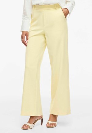 Femme portant un pantalon large jaune pâle, un chemisier blanc et des sandales à talons ouvertes beige, debout avec une main dans la poche.