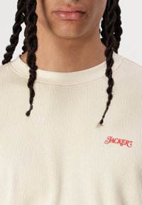 Cream ribbed sweatshirt featuring a round neckline and red embroidered logo on the chest. Accents include contrasting red stitching.