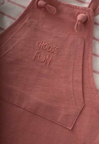 Los overoles de algodón rosa presentan un bordado que dice "Elige la Diversión" en el bolsillo, con tirantes ajustables y un acabado texturizado. Fondo con rayas visible.