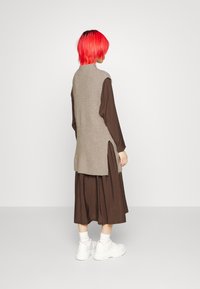 Gilet lavorato a maglia beige con un collo alto, fessure laterali, indossato sopra un vestito marrone; il modello indossa delle sneakers bianche e calzini bianchi.