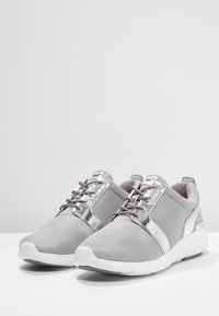 Szare buty sportowe wykonane z teksturowanej tkaniny z metalicznymi akcentami, z białymi podeszwami i szarymi sznurowadłami, zaprojektowane z myślą o komforcie i elastyczności.