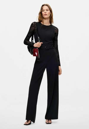 Mujer que lleva un mono negro con cinturón y mangas largas transparentes, sosteniendo un bolso negro y rojo, posando frente a un fondo blanco liso.