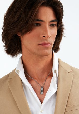 Silverkedja med ett rektangulärt hänge med texturerad yta, bärs över en vit skjorta och en beige blazer.