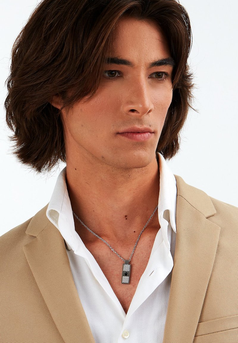 Sølv kæderekke med en rektangulær vedhæng med en struktureret overflade, båret over en hvid skjorte og en beige blazer.