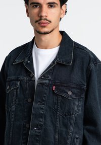 Giacca in denim scuro con un design classico, con accenti di bottoni metallici e tasche frontali. Indossata sopra una camicia bianca.